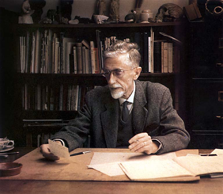 A color photograph of M.C. Escher lookit at some papers on his desk.